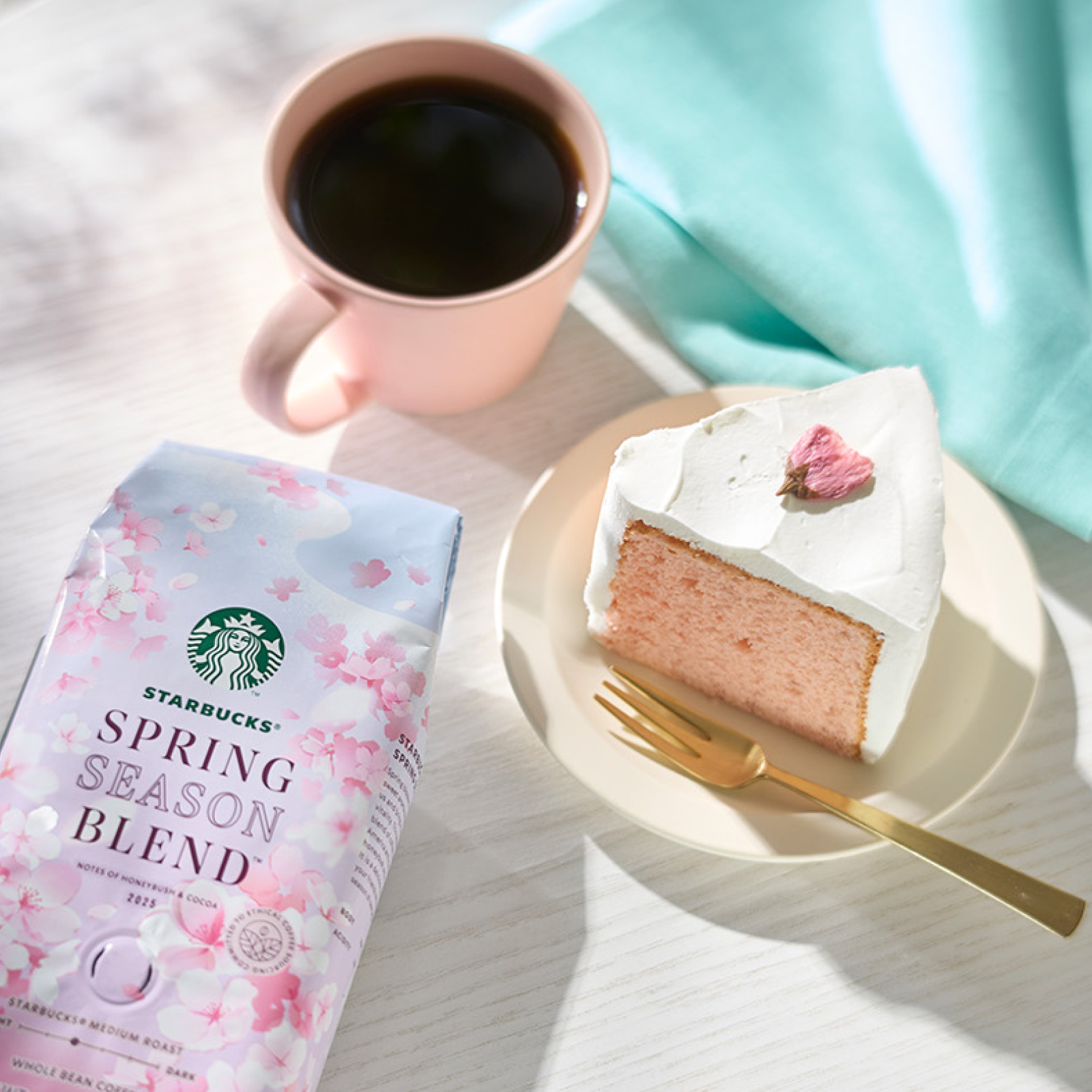 カフェテーブルに置いたスターバックス® スプリング シーズン ブレンド®のパッケージ、桜シフォンケーキと、ピンクのマグに注がれたコーヒー